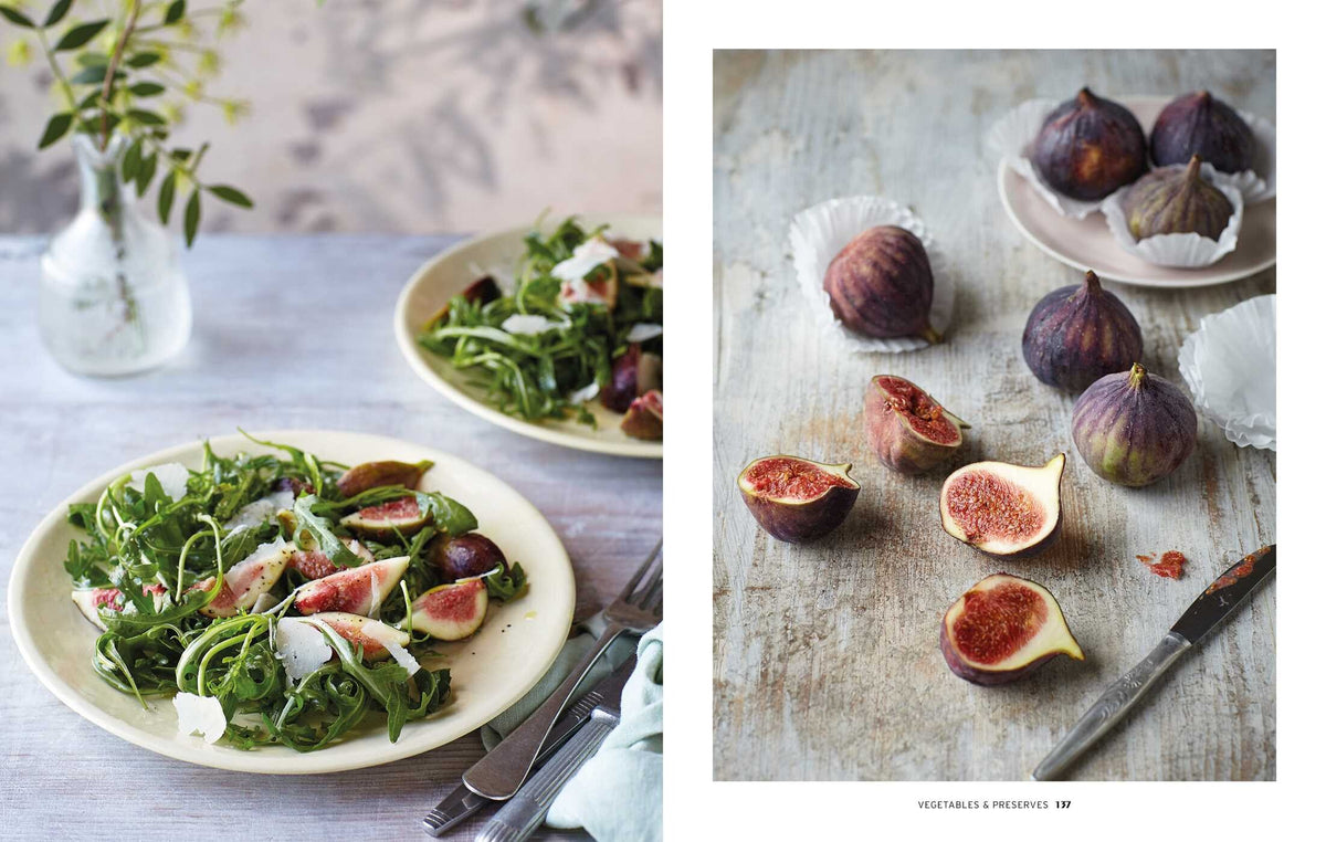 Two plates of arugula and fig salad on the left—an elegant nod to regional Italian cuisine inspired by Cucina Di Amalfi By Ursula Ferrigno from Simon & Schuster—while whole and halved figs with a knife and paper cups rest on the right, all arranged on rustic surfaces.