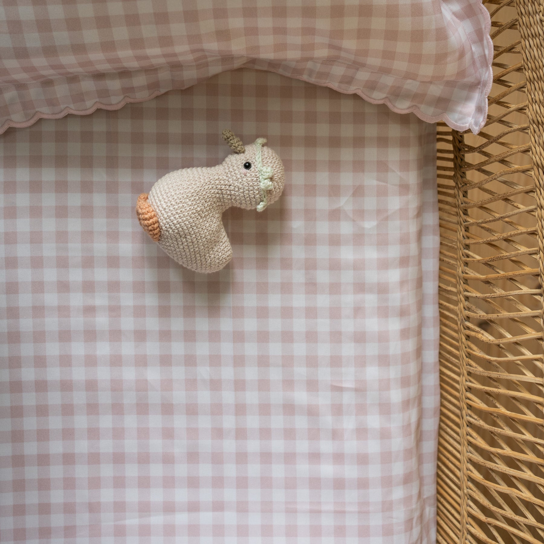 A small crochet animal toy rests on the Gooselings Picnic Gingham Pink Crib Sheet, which features an extra soft, OEKO-TEX certified fabric in a pink and white gingham pattern. A wicker frame appears along the edge.