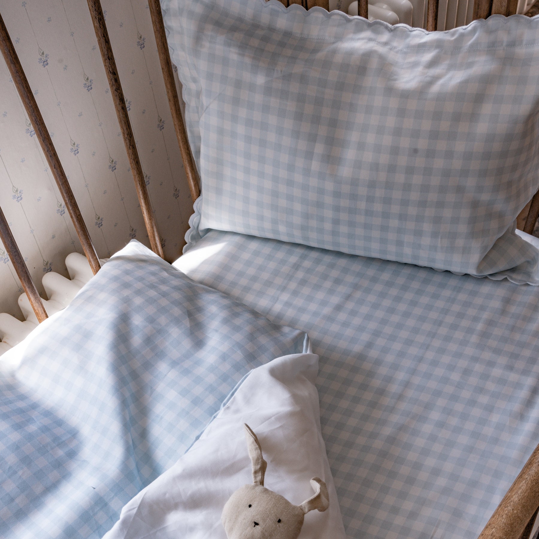 The Gooselings Picnic Gingham Blue Toddler Duvet, with its light blue and white checks, plus a matching pillow and soft rabbit toy on a white blanket, creates a cozy space—perfect before transitioning to a big-kid bed.