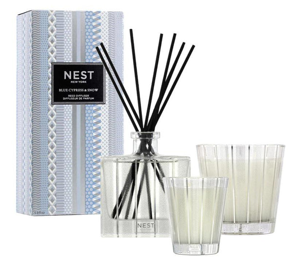 The NEST Blue Cypress & Snow Collection by Nest includes a fragrance set with a reed diffuser featuring black sticks, two white candles in glass holders, and a patterned box labeled "Blue Cypress & Snow," enriched with notes of Smoked Vanilla Bean.