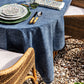A round table featuring the Juliska Blue Denim Tablecloth is set with patterned plates, bamboo-handle cutlery, and napkins. Classic wicker chairs surround the table, which sits atop a gravel surface.