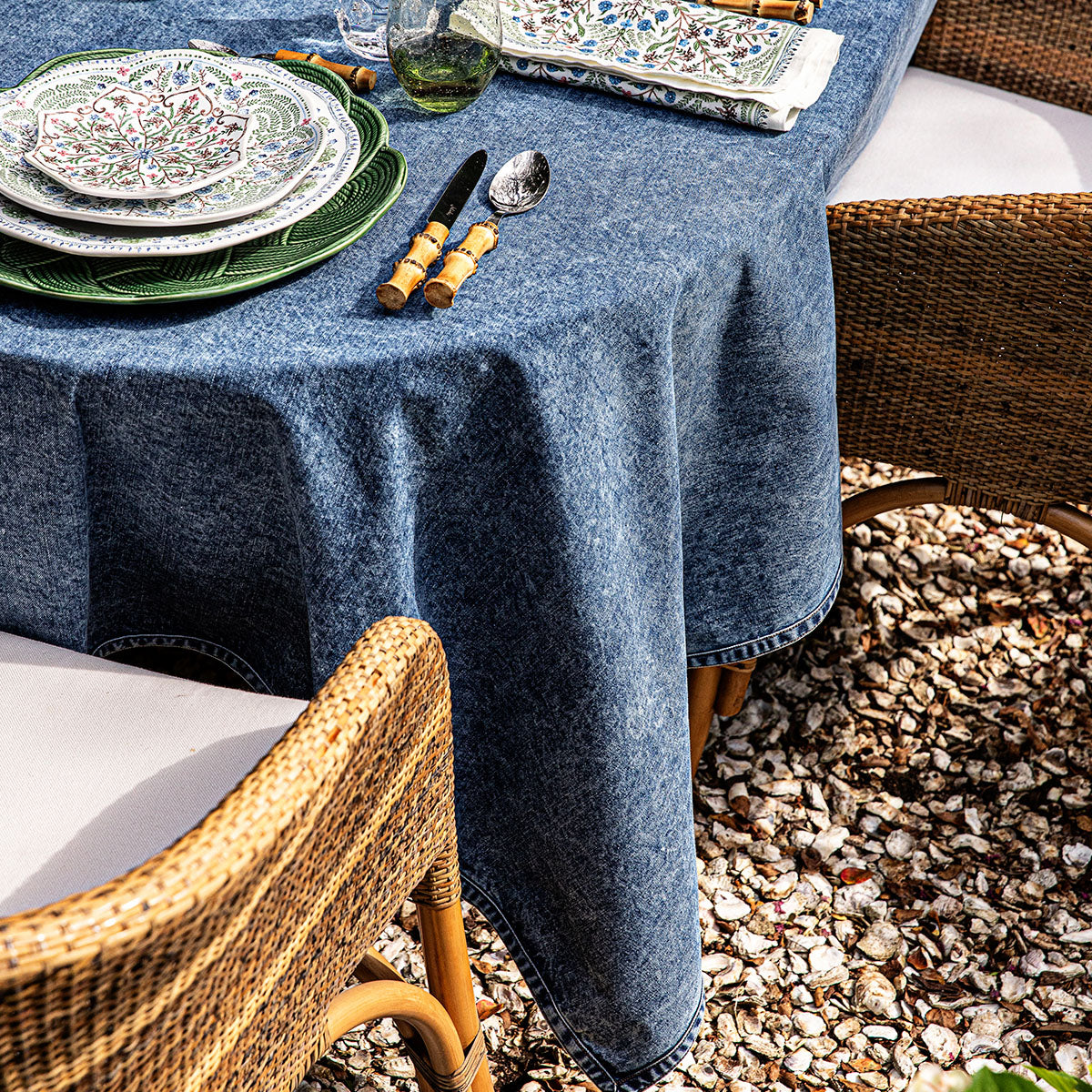 A round table featuring the Juliska Blue Denim Tablecloth is set with patterned plates, bamboo-handle cutlery, and napkins. Classic wicker chairs surround the table, which sits atop a gravel surface.