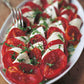 Sliced tomatoes and mozzarella artfully arranged on a white plate, topped with chopped fresh basil and a drizzle of olive oil, capture the simple home cooking traditions featured in Barefoot Contessa Back To Basics by Penguin Random House. A fork rests at the side.