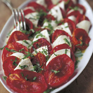 Sliced tomatoes and mozzarella artfully arranged on a white plate, topped with chopped fresh basil and a drizzle of olive oil, capture the simple home cooking traditions featured in Barefoot Contessa Back To Basics by Penguin Random House. A fork rests at the side.