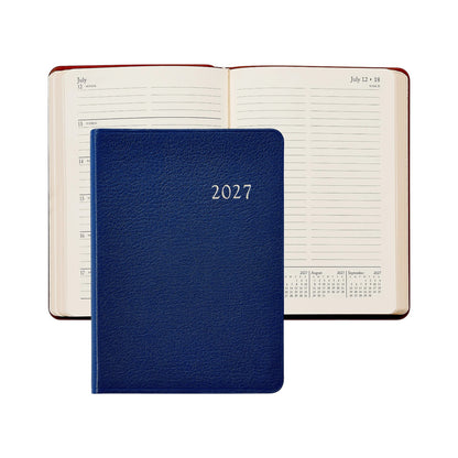 A closed Graphic Image 2027 Daily Journal in blue rests atop an open diary displaying a dated weekly layout alongside a small calendar.