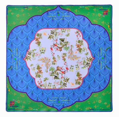 The Oh My Mahjong Mahjong Mat Collection features a square, non-slip mat with a blue scalloped pattern, green corners, and an Asian-inspired central design of trees, houses, and birds.