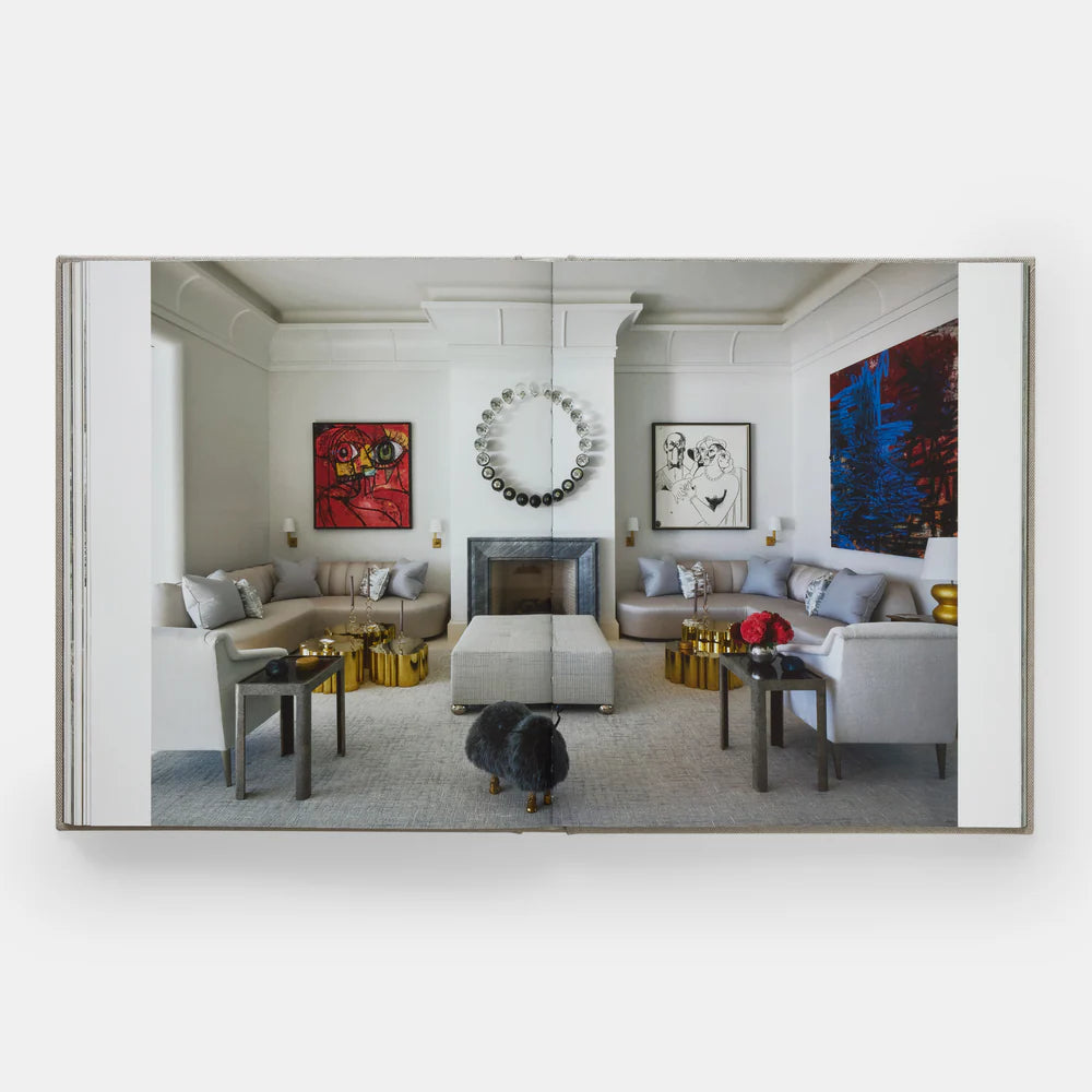 Open book of "Interiors" by Phaidon shows a modern living room with gray furniture, colorful abstract art, gold accents, and a circular mirror above the fireplace—highlighting luxury-inspired interior design.