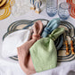 A table setting features a blue plate, bamboo-handled cutlery, Provence glassware, and three napkins—white, pink, green—each adorned with Juliska’s Provence Napkin Ring Collection for vibrant decor.