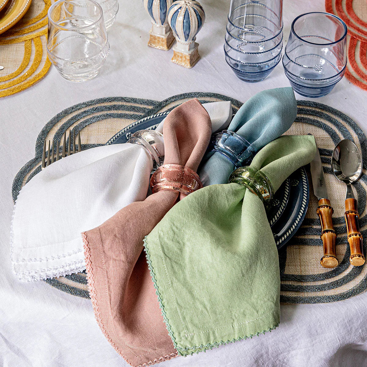A table setting features a blue plate, bamboo-handled cutlery, Provence glassware, and three napkins—white, pink, green—each adorned with Juliska’s Provence Napkin Ring Collection for vibrant decor.