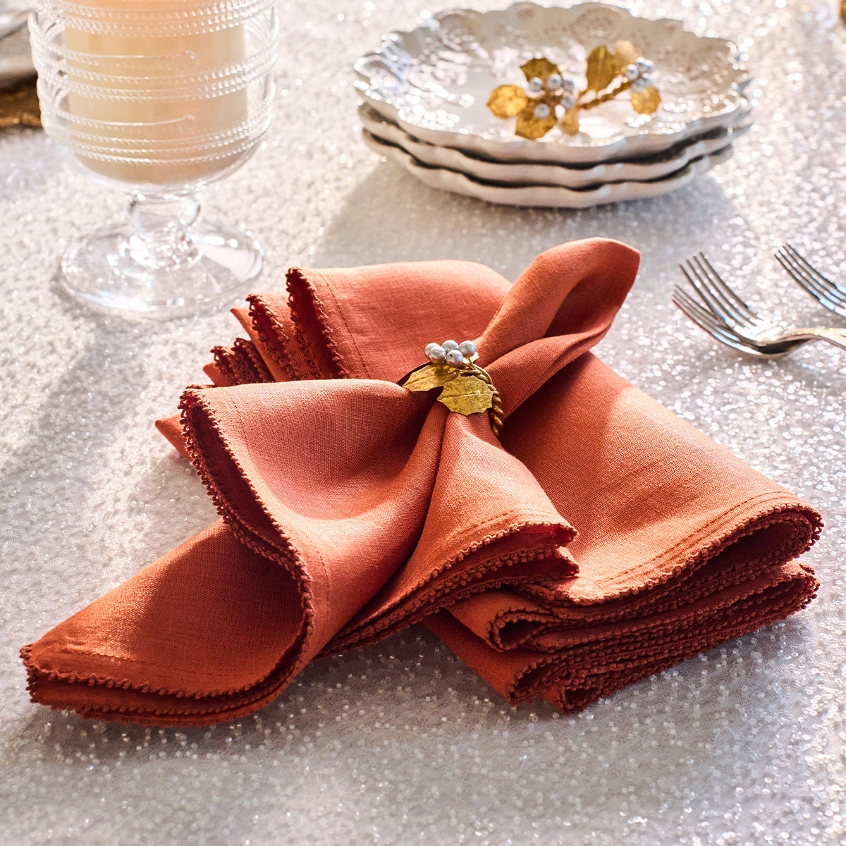 A Berry Scallop orange linen napkin adorned with a gold leaf and pearl napkin ring rests on a matching Berry Scallop woven linen tablecloth from Juliska, accompanied by Juliska plates, a candle, and two forks.