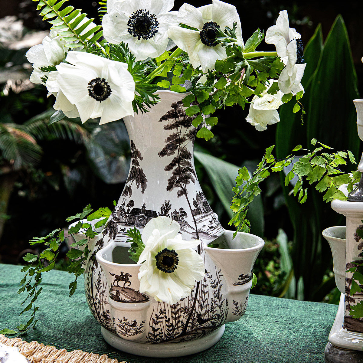The Juliska Flint Grey Country Estate Tulipiere Vase, featuring black and white landscape designs, holds white flowers with dark centers and greenery, enhancing the floral display on a green tablecloth outdoors.
