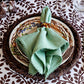 A Berry Trim Napkin from Juliska, featuring embroidered edging and a decorative ring, is displayed on a patterned plate with a woven charger on a leaf-print tablecloth.