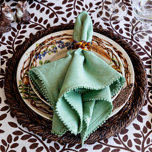 A Berry Trim Napkin from Juliska, featuring embroidered edging and a decorative ring, is displayed on a patterned plate with a woven charger on a leaf-print tablecloth.