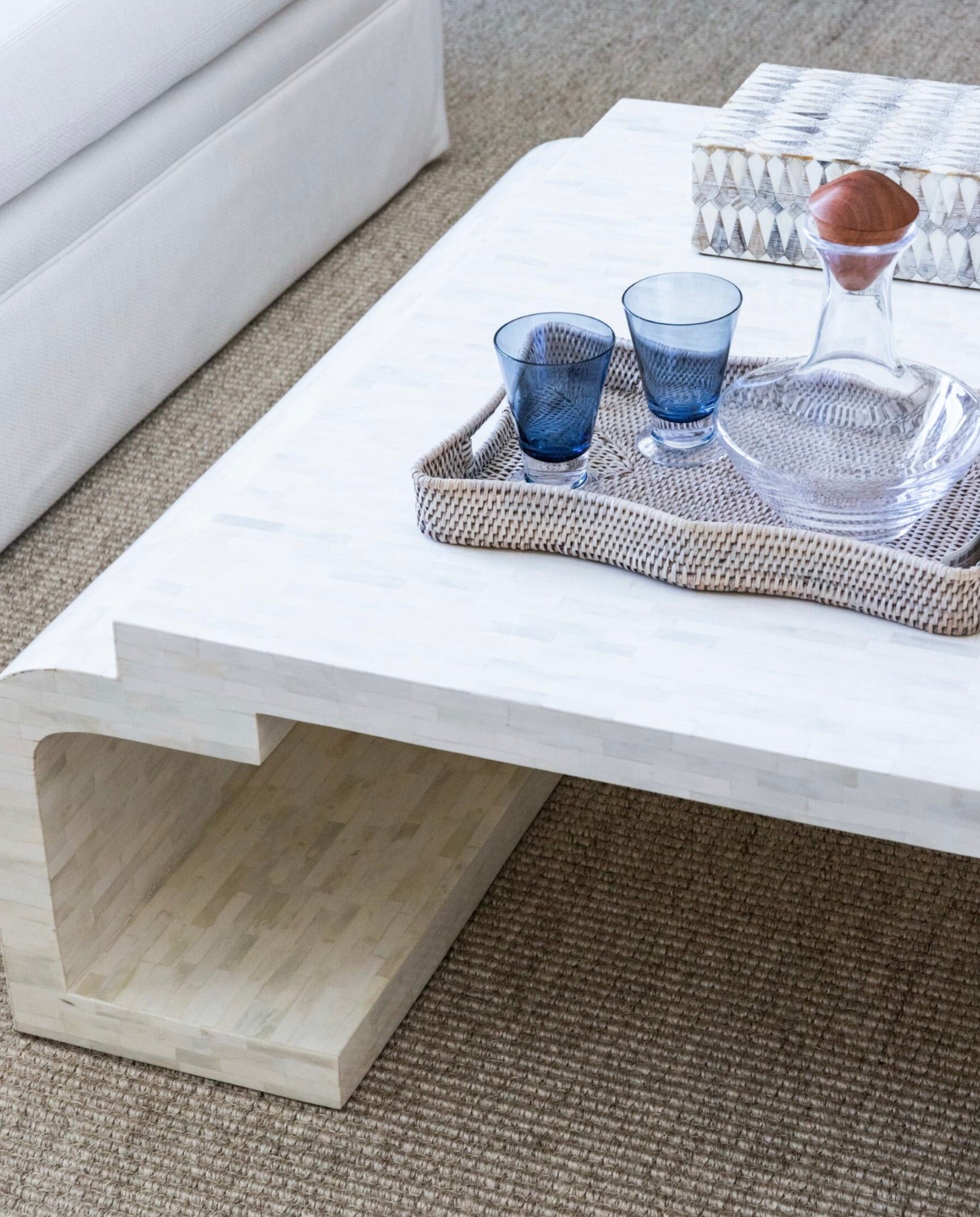 On a beige carpet next to a white sofa, there is a white coffee table adorned with the Opal Tray, Small by Made Goods, which holds two blue glasses and a crystal decanter.