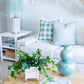 A light-filled living room with a white sofa, Hive Custom Pillows’ Small Check Blue Pillow (down-filled, 22" x 22"), a side table with lamp, round wooden coffee table, green plant, stacked books, and blue ceramic vases.