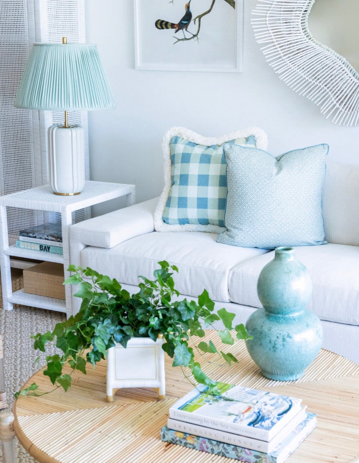 A light-filled living room with a white sofa, Hive Custom Pillows’ Small Check Blue Pillow (down-filled, 22" x 22"), a side table with lamp, round wooden coffee table, green plant, stacked books, and blue ceramic vases.
