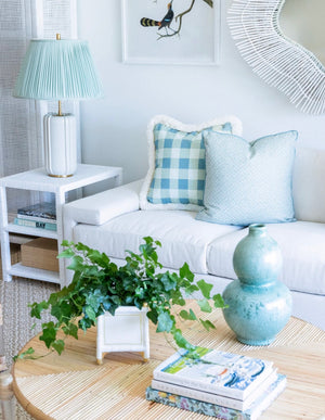 In the living room, a white sofa is beautifully adorned with blue and white cushions. On the coffee table sits a lush plant in a Green Gourd Vase from the Van Cleve Collection, complemented by a glass lamp. A decorative mirror on the wall enhances the room's elegance.