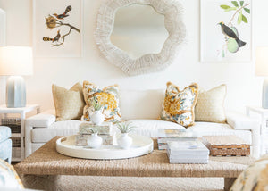 Modern living room with a white Denby Sofa by Hickory Chair adorned with floral cushions featuring Spring-Down seat cushions, a large circular mirror above, bird-themed wall art, and a coffee table displaying decor items, books, and a woven tray.