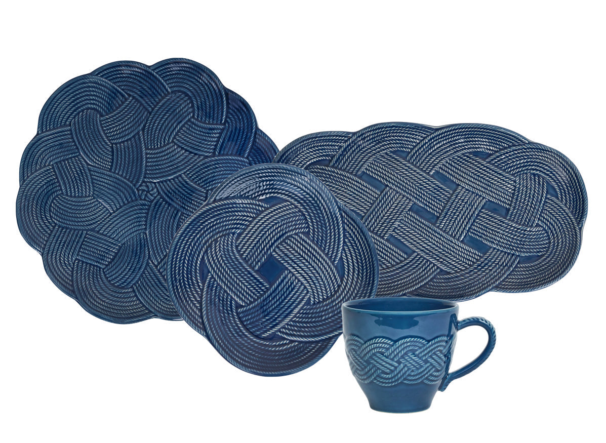 Four blue woven placemats from Juliska’s Le Panier Mystic Knot Denim Dinnerware and Serveware Collection, featuring intricate mystic knot designs, are displayed behind a matching blue Juliska mug adorned with a similar knot motif, evoking the artful elegance of a large charger or serving platter.