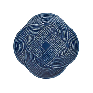 A round charger plate from the Juliska Le Panier Mystic Knot Denim Dinnerware and Serveware Collection, featuring a denim blue glaze and a white braided rope pattern in a symmetrical mystic knot design.