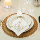 A refined table setting featuring white plates on a gold placemat, complemented by a folded Juliska Heidi Gold/Silver Embroidered Napkin held in a gold ring adorned with an Alpine floral motif, with glassware in the background.