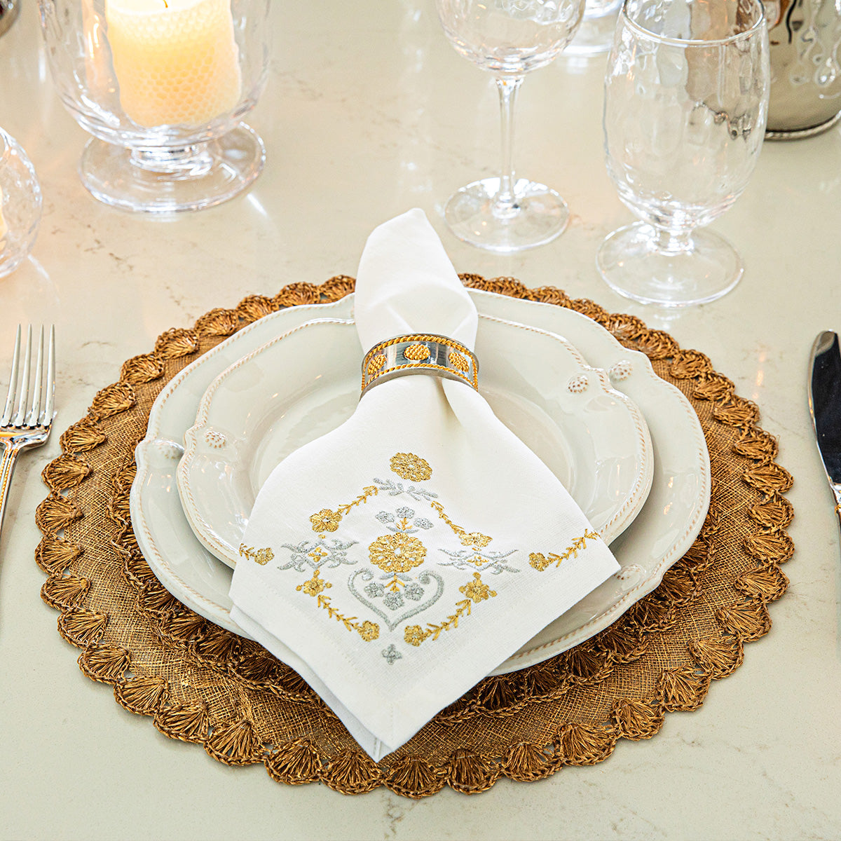 A refined table setting featuring white plates on a gold placemat, complemented by a folded Juliska Heidi Gold/Silver Embroidered Napkin held in a gold ring adorned with an Alpine floral motif, with glassware in the background.
