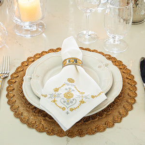 A refined table setting featuring white plates on a gold placemat, complemented by a folded Juliska Heidi Gold/Silver Embroidered Napkin held in a gold ring adorned with an Alpine floral motif, with glassware in the background.