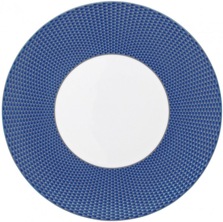 The Tresor Blue Dinnerware Collection by Raynaud features a round white porcelain plate with a wide, blue textured rim and small checkered pattern, adding an elegant touch to any table setting.