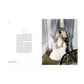 A woman in a white gown adorned with floral details stands at a window, holding open gray curtains in an elegant, luminous interior with a sweeping staircase—a scene that beautifully captures the timeless sophistication and delicate allure featured in Rizzoli’s *Dior in Bloom*.