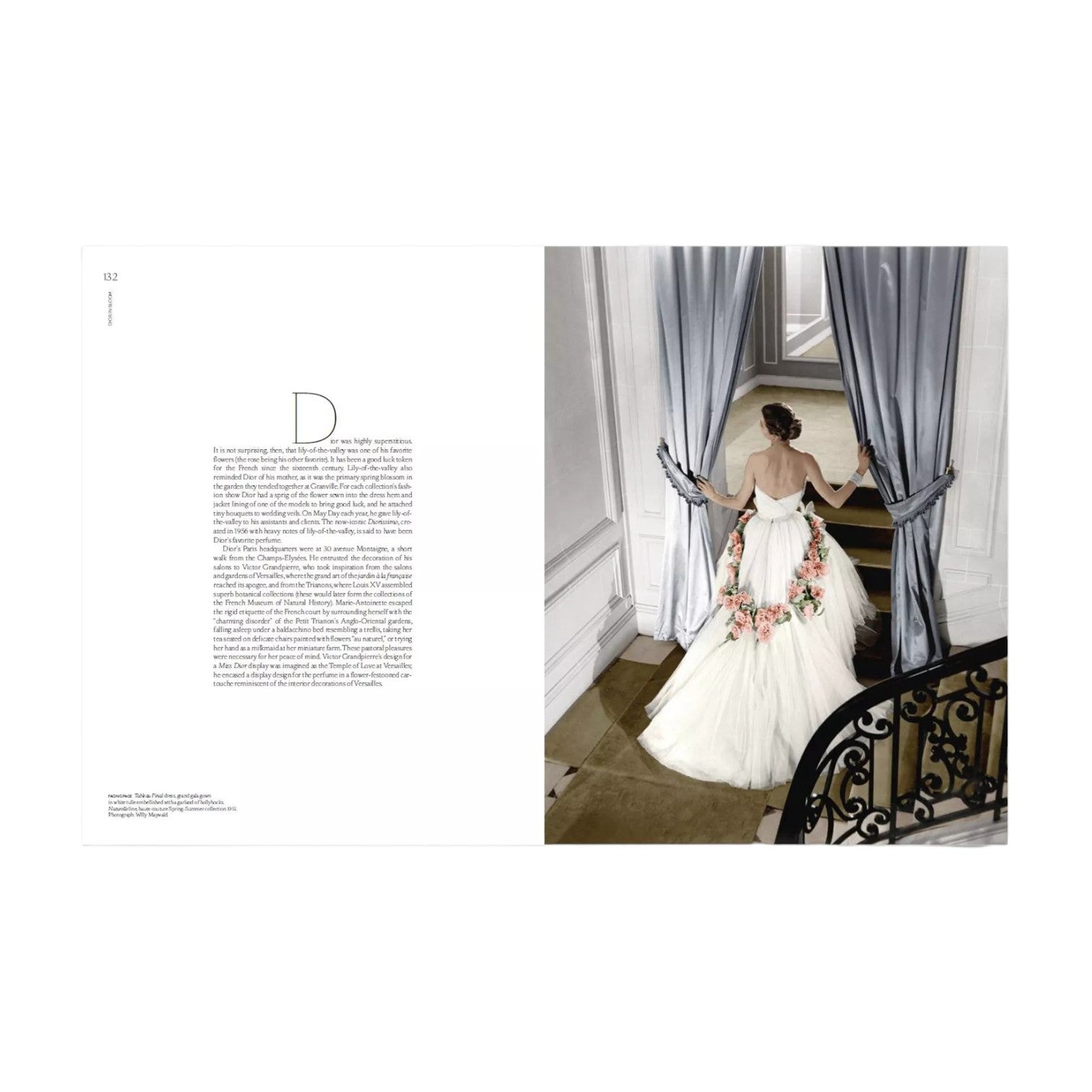 A woman in a white gown adorned with floral details stands at a window, holding open gray curtains in an elegant, luminous interior with a sweeping staircase—a scene that beautifully captures the timeless sophistication and delicate allure featured in Rizzoli’s *Dior in Bloom*.
