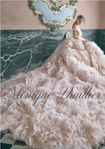 A woman wearing a strapless, ruffled blush gown—evoking the elegance of Monique Lhuillier as featured in Penguin Random House’s collection—sits by a green marble wall beneath a decorative mirror. The words “Monique Lhuillier” appear in script across the image.
