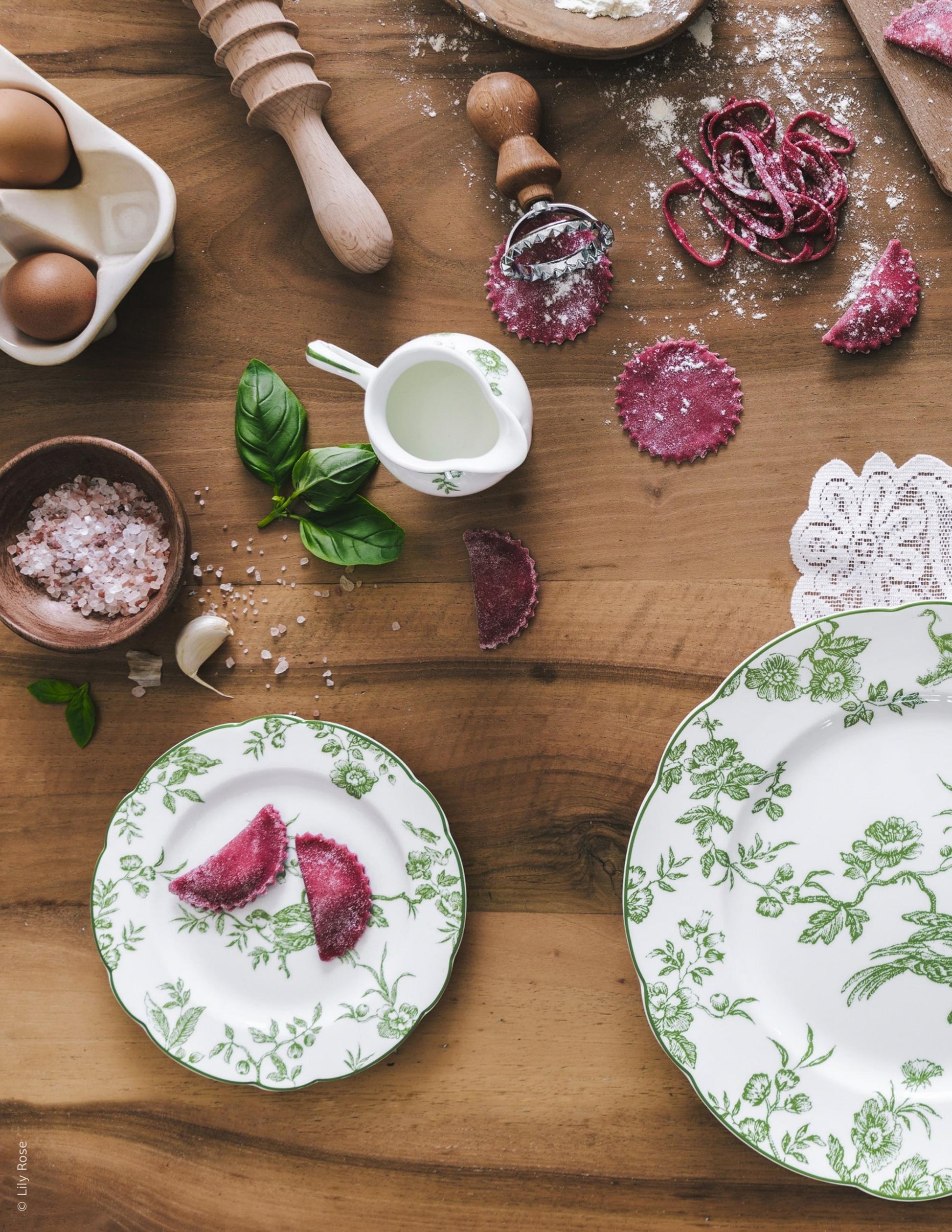 A wooden table with eggs, salt, flour, pasta tools, purple ravioli, a small jug of milk, fresh basil, and Bernardaud’s Albertine Collection green-patterned plates with scalloped edges arranged neatly.