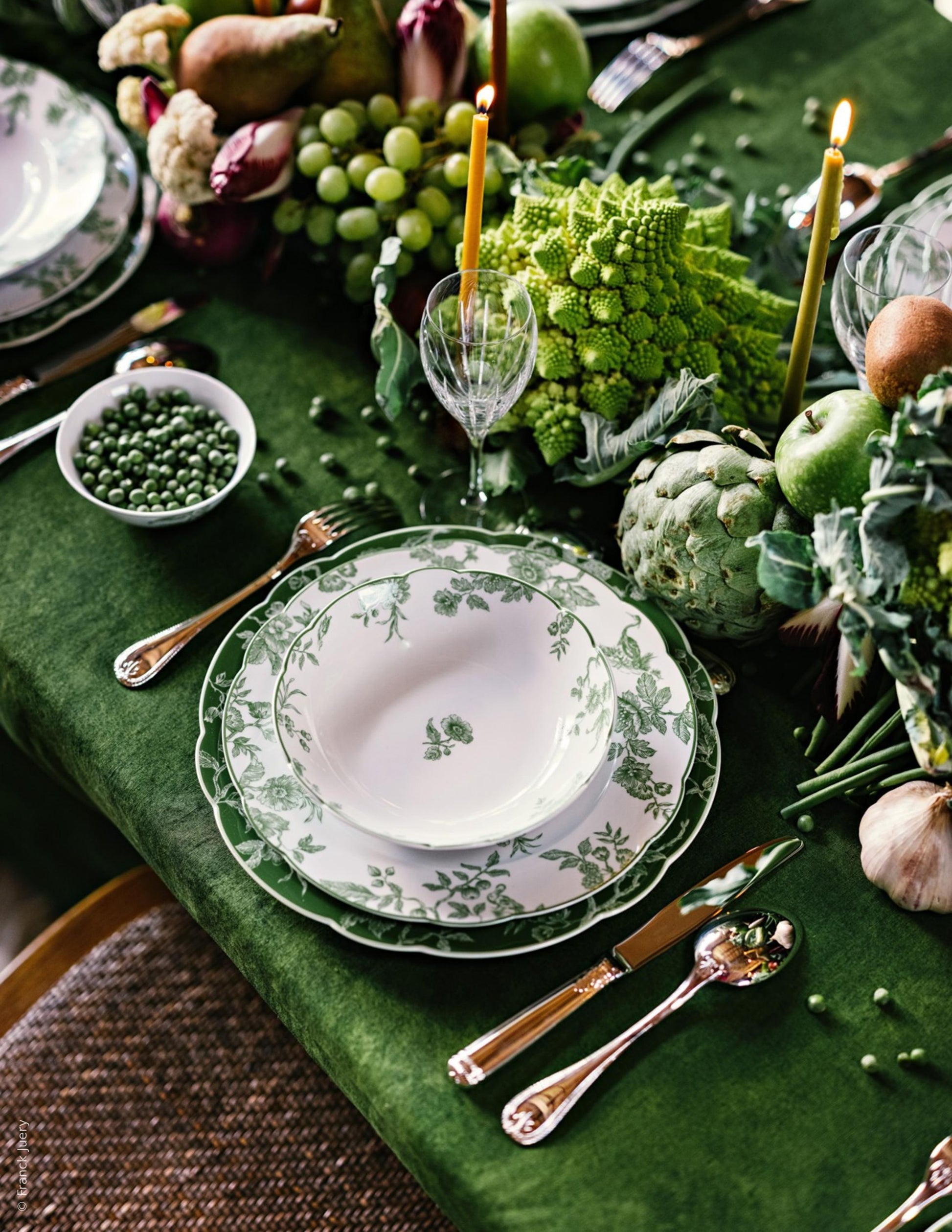 The Albertine Collection by Bernardaud, with its scalloped-edge plates, silver cutlery, candles, wine glasses, peas, and fresh vegetables on a green tablecloth, creates a poetic green-themed table decor.