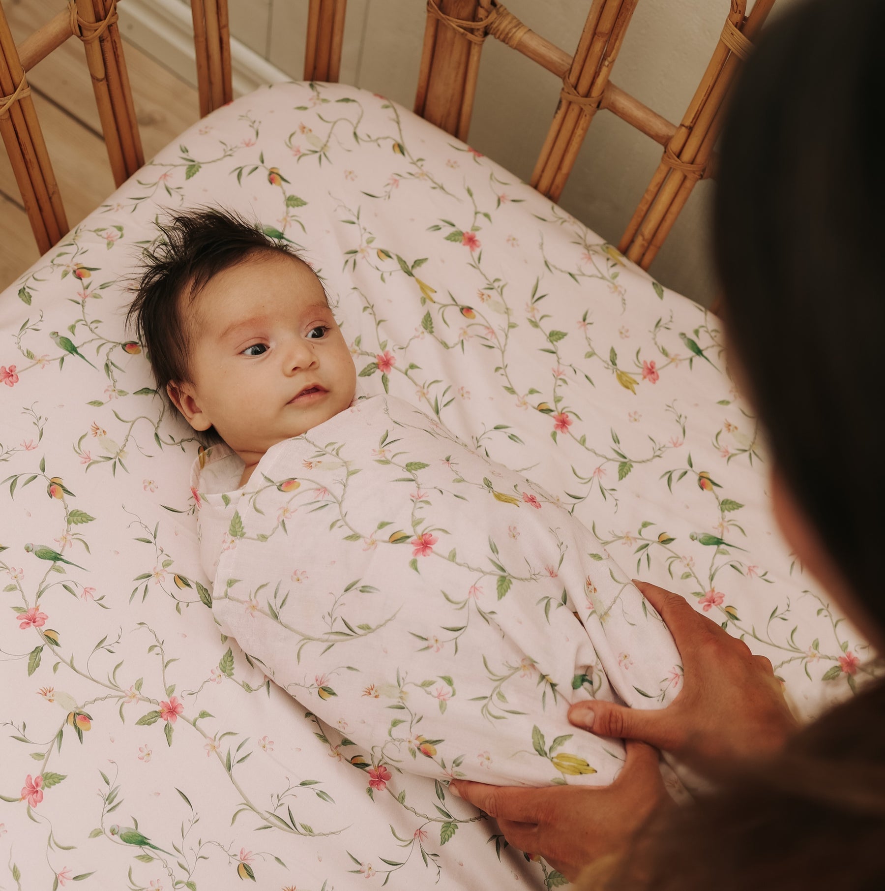 A baby is swaddled in the Gooselings Dreaming of Paradise Swaddle, lying on a matching crib sheet in the nursery as an adult’s hands gently hold them.