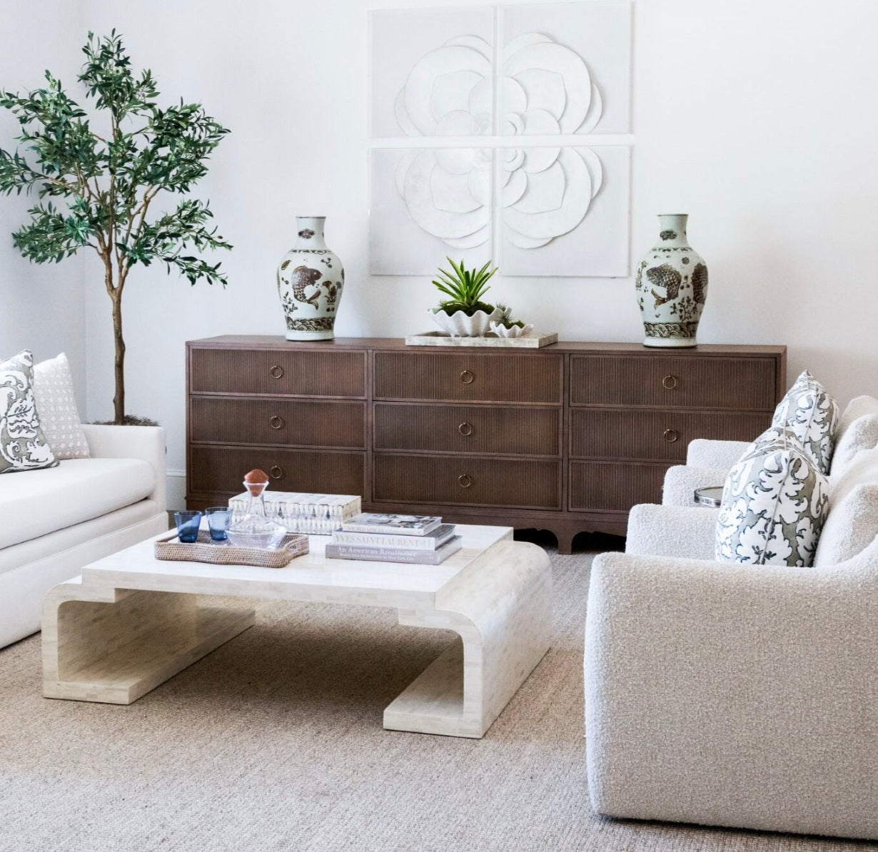 A modern living room featuring a white couch and two white armchairs around a marble coffee table. The space is enhanced by the Century Furniture Reeded Dresser, showcasing a reeded case front in charcoal finish, alongside a wooden cabinet adorned with decorative vases. A large plant in the corner completes the look.