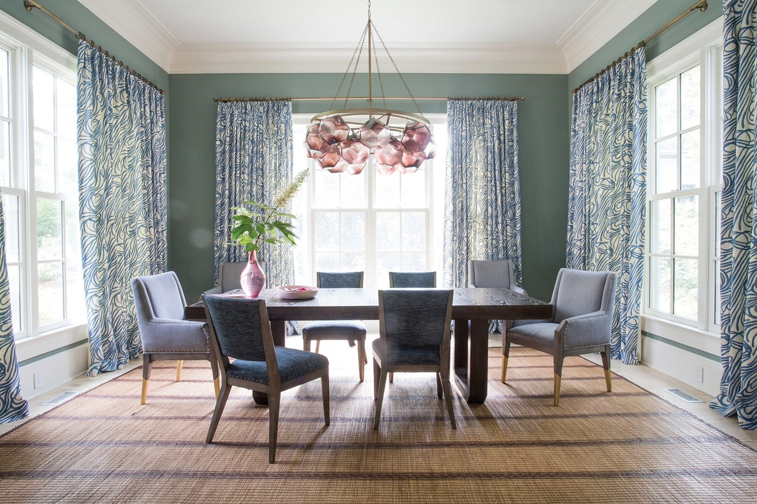 A dining room featuring the Hickory Chair Oliver Extension Dining Table with six gray chairs, a woven rug, blue patterned curtains, a pink chandelier, and a vase of greenery as the centerpiece.