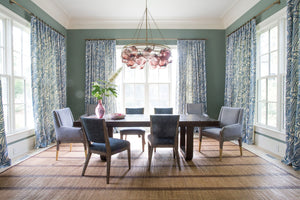 A dining room featuring the Hickory Chair Oliver Extension Dining Table with six gray chairs, a woven rug, blue patterned curtains, a pink chandelier, and a vase of greenery as the centerpiece.