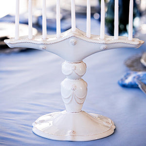 The Juliska Berry & Thread Classic Whitewash Menorah, with eight unlit candles, is displayed on a blue tablecloth.