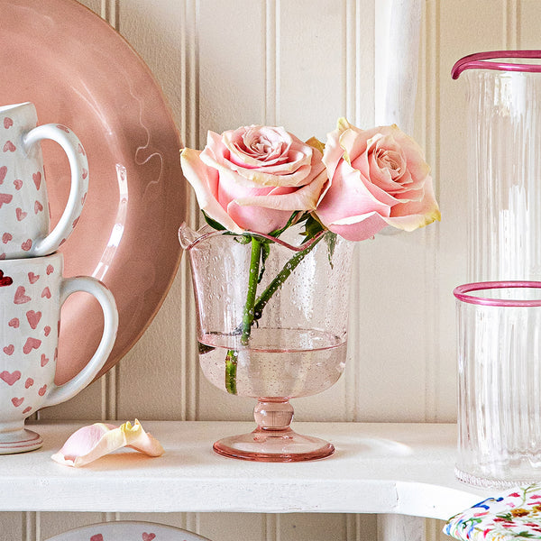 Two pink roses arranged in the Juliska Provence Vase, a whimsical pink mouth-blown glass piece by Juliska, are displayed on a shelf among heart-patterned mugs, a plate, and clear glasses with pink rims.