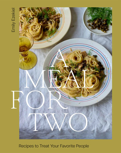 Two plates of pasta with herbs and capers, paired with two glasses of white wine on a white tablecloth. Text on image reads: “A Meal For Two” by Chronicle Books. Ideal for couples or duos, this cookbook offers simple yet delicious recipes to make weekday meals feel extra special.