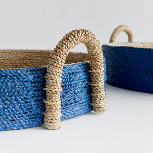 Two round handwoven baskets from the Tiana Blue Seagrass Round Basket Collection by Napa Home & Garden, featuring blue bodies and natural handles, are displayed on a white surface against a plain background.