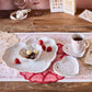 A wooden table set with Juliska’s Berry & Thread Whitewash Heart Serveware Collection, ruffled rim plates, a mug, chocolates, strawberries, pink accents, and greeting cards—perfect for a romantic or Valentine’s Day celebration.