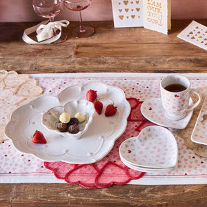 A wooden table set with Juliska’s Berry & Thread Whitewash Heart Serveware Collection, ruffled rim plates, a mug, chocolates, strawberries, pink accents, and greeting cards—perfect for a romantic or Valentine’s Day celebration.