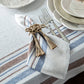 A folded napkin from Juliska’s Danish Stripe Table Linen Collection, held by a knotted rope ring, sits beside two forks on a matching linen tablecloth, showcasing minimalist Scandinavian elegance.
