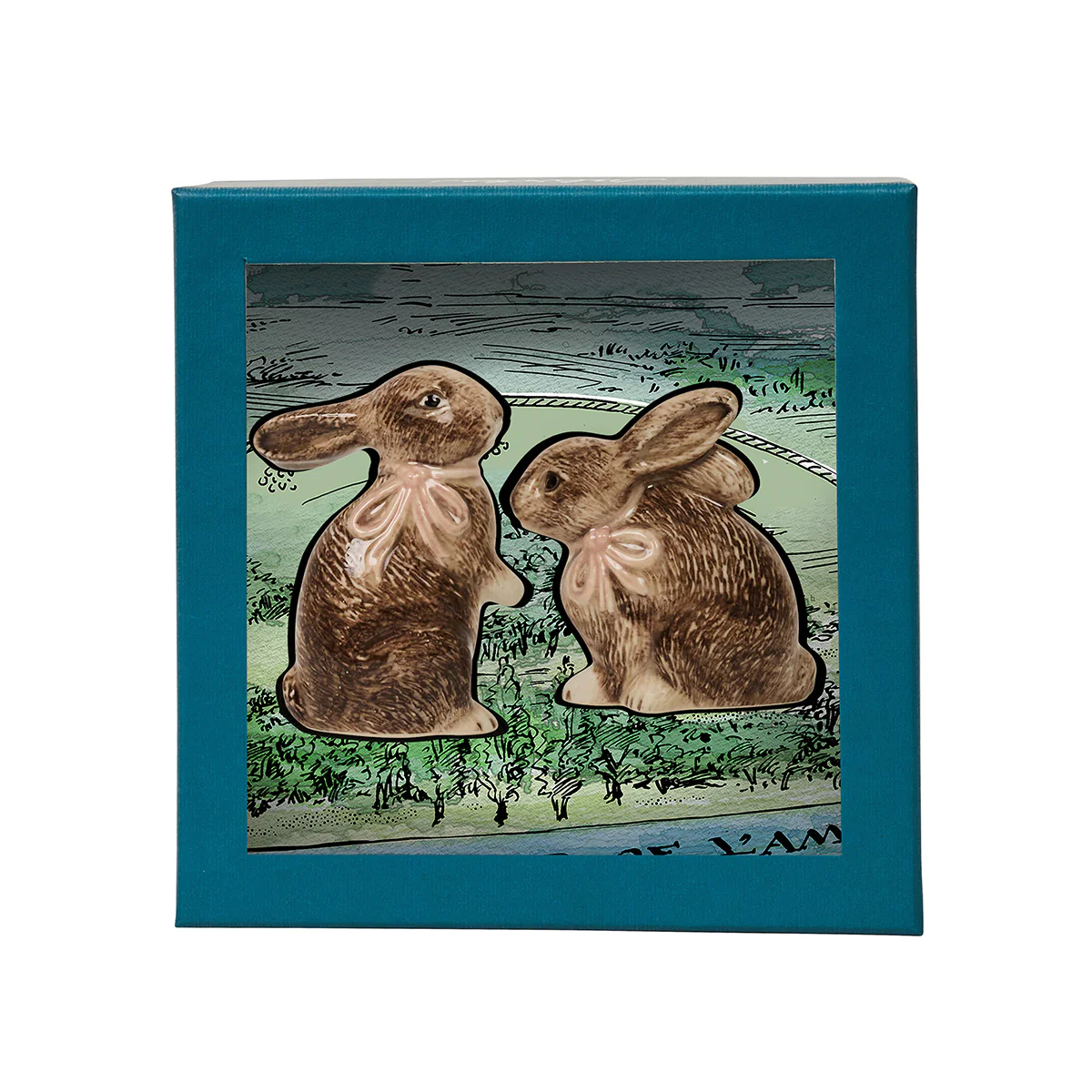 The Juliska Clever Creatures Bunny Salt and Pepper Set features two hand-painted ceramic bunnies with pink bows, presented in a gift box with blue edging and a green illustrated background.