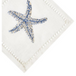 A white cloth napkin from Haute Home’s Starfish Coaster & Bath Collection, crafted from Italian linen, showcases a blue and white embroidered starfish in one corner and decorative stitching along the edges—an elegant accent that pairs beautifully with other luxurious linen coasters and refined home décor pieces.