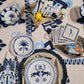 A table set with Johanna Ortiz’s Casa Deep Navy Dinnerware Collection, cutlery, napkins, lemons in a small bowl, and packaged cheese on a decorative tablecloth highlights artisanal heritage.