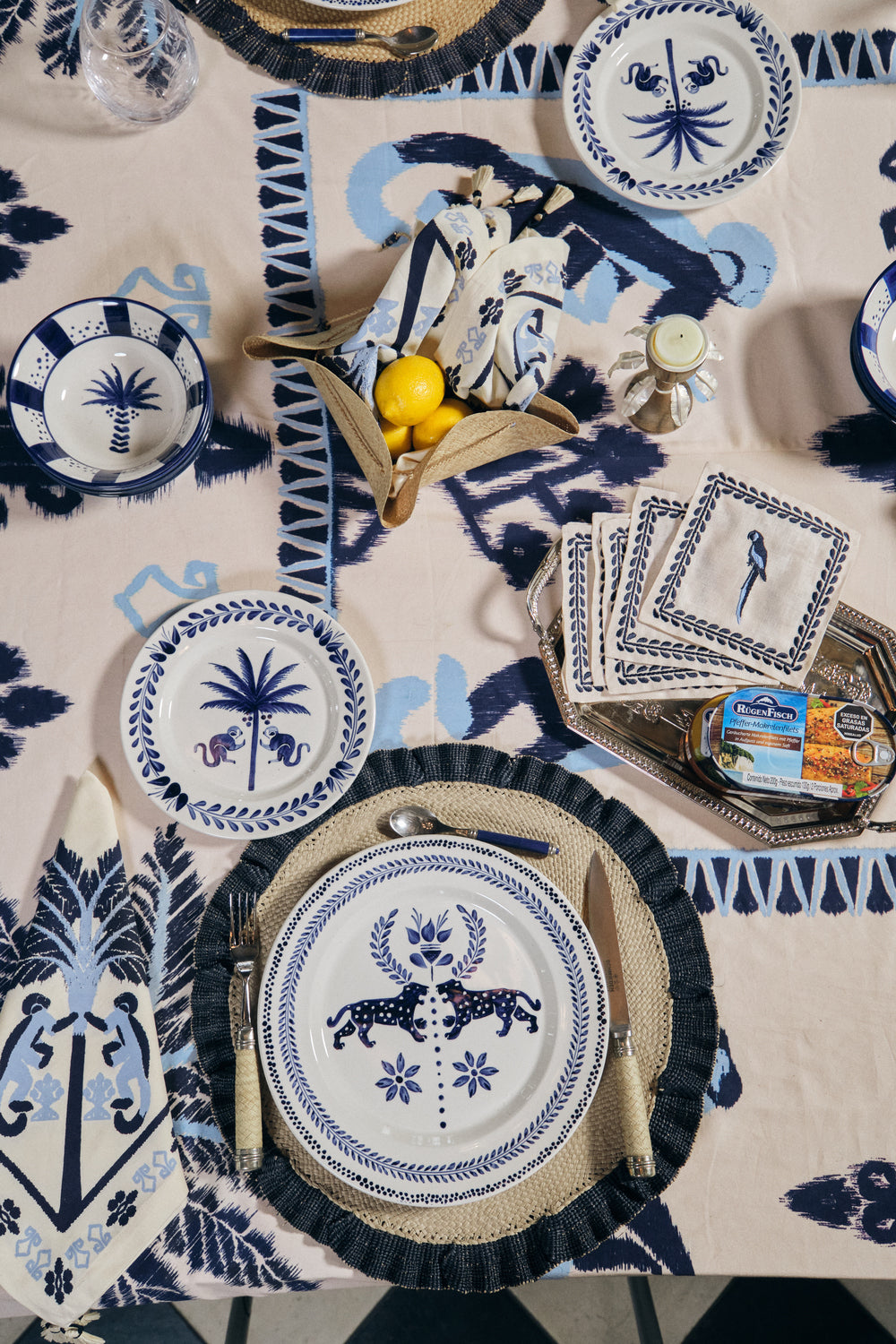 A table set with Johanna Ortiz’s Casa Deep Navy Dinnerware Collection, cutlery, napkins, lemons in a small bowl, and packaged cheese on a decorative tablecloth highlights artisanal heritage.