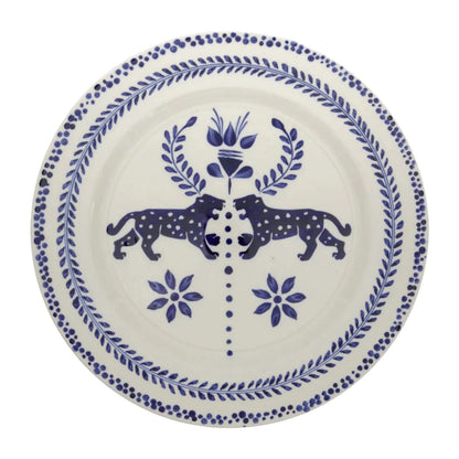 The Casa Deep Navy Dinnerware Collection by Johanna Ortiz features white ceramic plates with blue motifs—two mirrored spotted animals facing a central floral design—showcasing hand-painted artistry and the heritage of Carmen de Viboral clay.