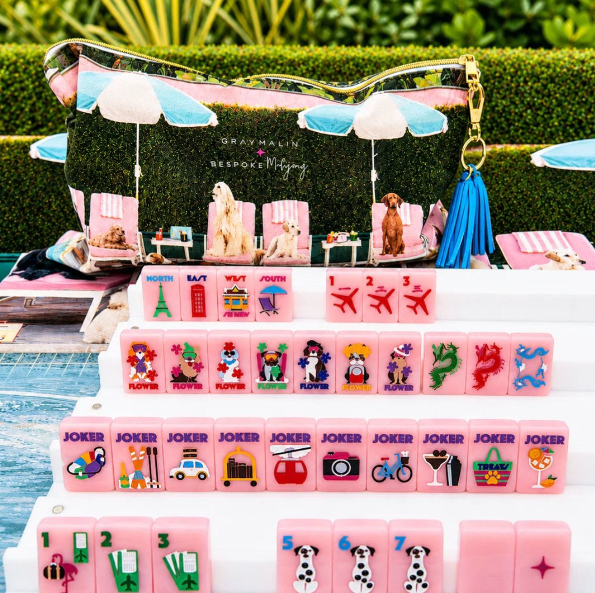 Bespoke x Gray Malin Mahjong Collection set with pink tiles featuring travel icons and dogs, arranged on racks in front of a zippered pouch printed with a beach scene.